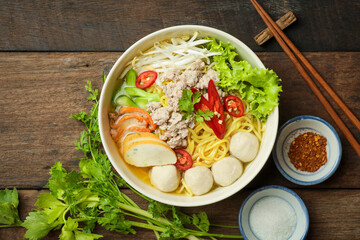 Delicious bowl of Asian noodle soup with fish balls, minced pork, fresh vegetables, and spicy red chili, served on a rustic wooden table with chopsticks.
