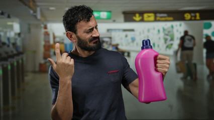 Hispanic man with beard confusedly examines a pink detergent bottle in busy airport terminal,...