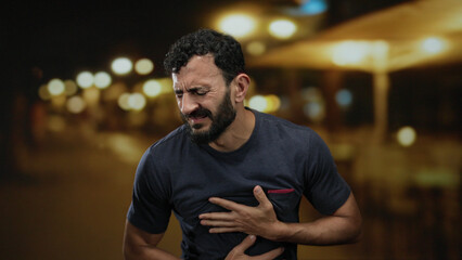 Obraz premium Man with beard showing expression of discomfort on a dimly lit street with bokeh lights outdoors at night.