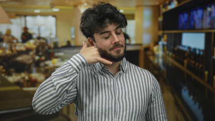 Fototapeta premium Young man with beard makes call gesture in lively restaurant ambiance suggesting friendly communication in a relaxed indoor setting with coffee and hispanic cultural nuances.