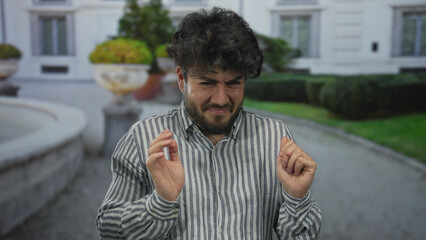 Young man with beard makes a gesture of disgust outdoors on a street with greenery and...