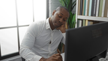 Young man in office wearing white shirt looking thoughtful at computer while resting his head on...