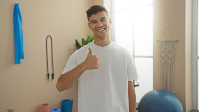Young man smiling in gym giving thumbs up wearing white shirt with gym equipment in background fitness center healthy lifestyle positive mood hispanic athlete indoors