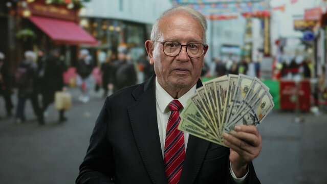 Senior man holding us dollars outdoors on bustling street expressing wealth and business success in urban setting with colorful shops and vibrant atmosphere - Powered by Adobe