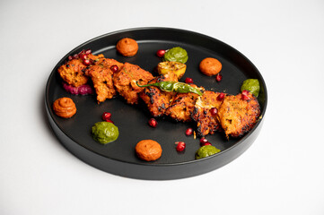  Classic chicken tikka served on a black plate with vibrant green and orange chutneys, garnished with pomegranate seeds, placed on a white background — a flavorful and authentic Indian preparation.