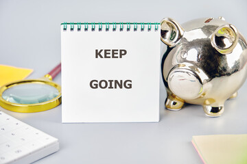 A motivational note with the words keep going is placed next to a shiny piggy bank on a workspace....