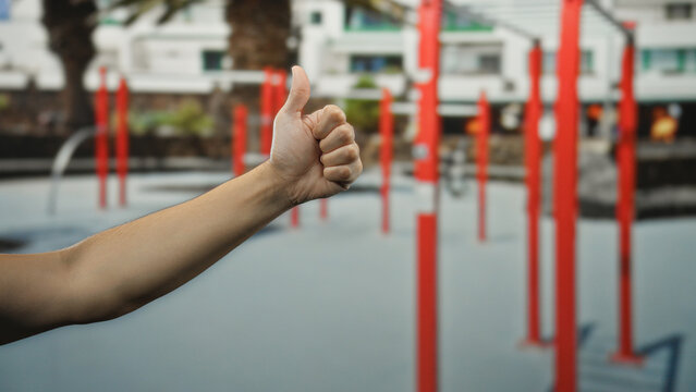 Man's hand giving thumbs up at outdoor gym with red structures in sports center background, symbolizing approval and fitness motivation in a lively environment.