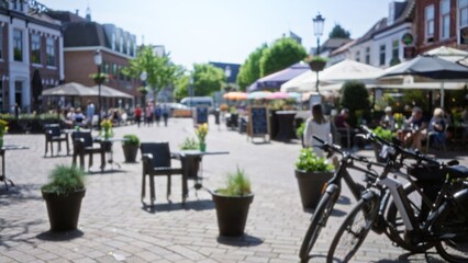 Blurred european town square with people enjoying outdoor cafes, vibrant flowers, bicycles, and historic architecture under a clear blue sky creates a picturesque urban ambiance.