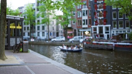 Fototapeta premium Blurred view of people enjoying a boat ride on an amsterdam canal, highlighting the charming architecture and serene waterscape under soft evening light.
