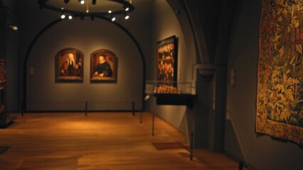 Blurred view inside an art gallery with paintings and tapestry, featuring dim lighting and wooden floors, creating a calm and contemplative atmosphere for visitors.