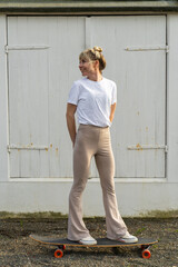 Self-confident woman in casual urban clothing standing on skateboard, posing at white village gate....