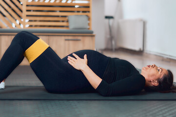 Pregnant woman lying down on yoga mat using resistance band during workout and doing breath work, preparing for childbirth  