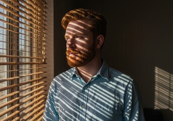 Red haired man stands by a window with sunlight streaming through the blinds