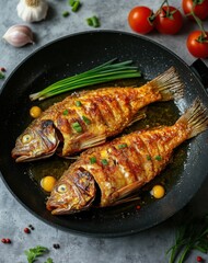 Top view of golden fried fish in a black pan with oil and green onions next to it on a grey concrete table