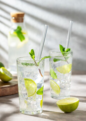 Two glasses mojito cocktail with fresh lime, mint and ice on light background with bottle and shadow.