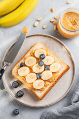Peanut butter toast with banana and blueberry on a plate on a light background with morning shadow.