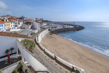 Sandy beach cove flanked by marina and mountain road on Terceira Island coastline. Aerial view of harbor entrance with shimmering Atlantic waters, stone breakwater and Mont Brasil hillside, Azores