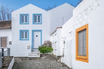 Charming White Houses with Blue and Orange Accents, Azores. Picturesque view of a bright white villa with distinctive blue window and door frames and an orange-framed window, set on a cobblestone path