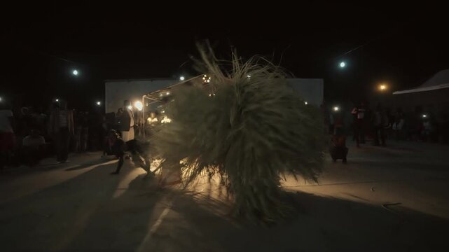 Senegalese Kumpo dancing at night around a ceremonial stick in Casamance &mdash; traditional costume made of straw spinning under spotlights, festive crowd filming the ritual celebration under magical light