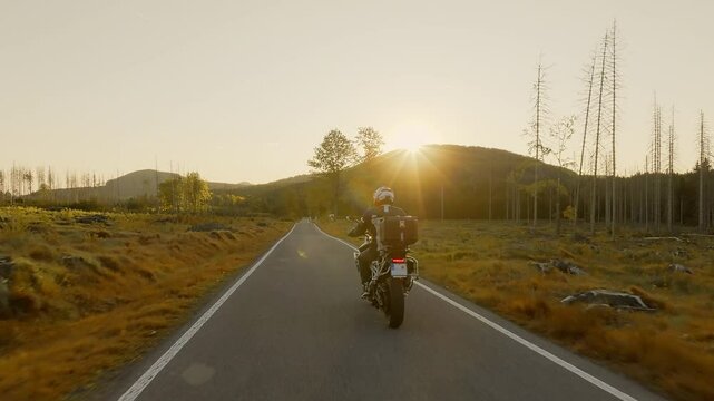 Driver riding motorcycle on empty asphalt road, spring mountains during sunset