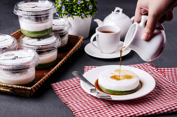 A hand pours brown sugar syrup over kue talam, a traditional green and white layered dessert on a plate, with several other desserts in plastic cups, a teapot, and a cup of tea in the background