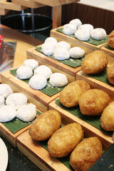 A variety of traditional snacks, including white powdered mochi-like balls and golden-fried oval pastries, are displayed on a tiered wooden buffet stand with banana leaf accents