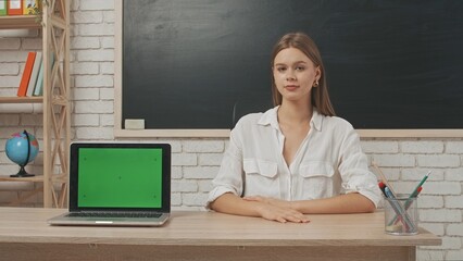 Young woman college teacher at desk in classroom in front of chalkboard explains lesson to students, laptop chroma key green screen. Education concept.
