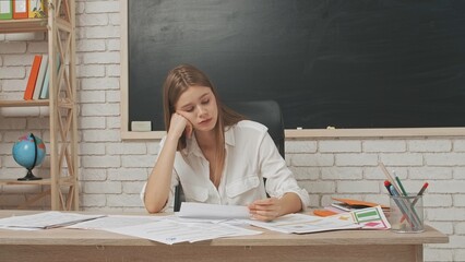 Young woman college teacher at desk in classroom in front of chalkboard checking homework, tires...