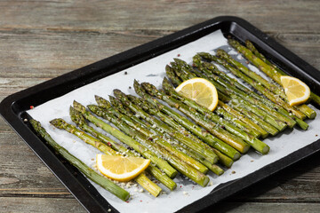 Baked green asparagus with lemon on baking tray and hard cheese on wooden surface