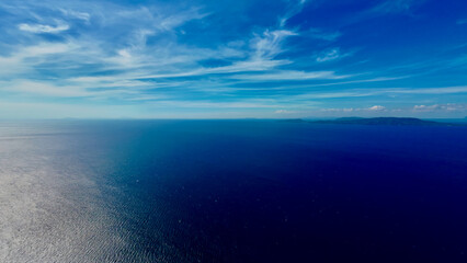 Obraz premium Aerial view of the sea surface and clouds above the horizon. Aerial view of the calm surface of the blue ocean, white clouds above the horizon and a small tropical island in the distance.