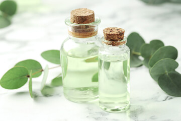 Bottles of essential oil and eucalyptus leaves on white marble table, closeup