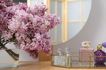 Beautiful lilac flowers in glass vase and perfumes on wooden table indoors, closeup