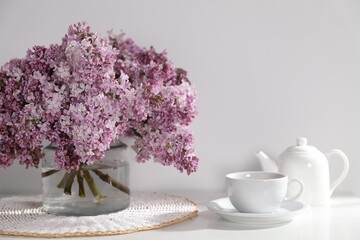 Beautiful lilac flowers in glass vase, cup of drink and teapot on table near white wall indoors