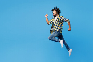 Full body side profile view young happy Indian man he wear yellow checkered shirt casual clothes jump high do winner gesture isolated on plain blue cyan background studio portrait. Lifestyle concept.