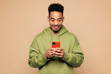 Young smiling man of African American ethnicity wear green hoodie casual clothes hold in hand use mobile cell phone isolated on plain pastel light beige background studio portrait. Lifestyle concept.
