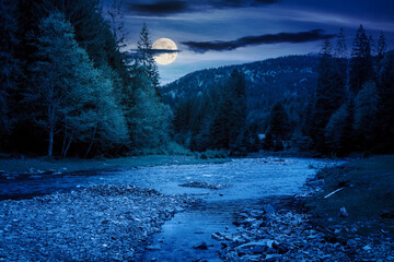 mountain landscape with river at night. mysterious scenery with stones in the water and forest on the shore in full moon light. starry sky with dark clouds