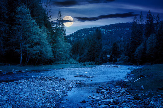 Fototapeta mountain landscape with river at night. mysterious scenery with stones in the water and forest on the shore in full moon light. starry sky with dark clouds