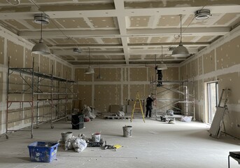 Interior space under construction shows workers on scaffolding and unfinished drywall