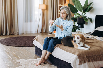 Senior woman using lancing device with beagle dog on bed at home