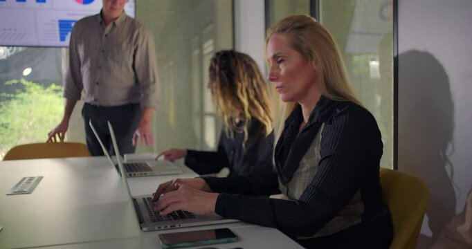 Focused blonde businesswoman typing on laptop while colleagues and manager engage in meeting discussion in well lit modern conference room