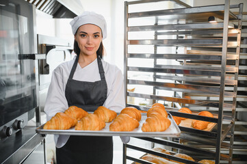 Baker woman holding tray of freshly baked croissants in bakery kitchen