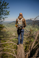 Naklejka premium Adventurous Hiker Exploring Naturpark Ammergauer Alpen: Lone Figure Standing On Rocky Outcrop Overlooking Majestic Mountain Ridges And Lush Green Valley Under A Clear Blue Sky In Scenic Bavaria