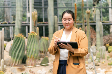 Asian female botanist using digital tablet and stylus to study and record cactus plant data inside...