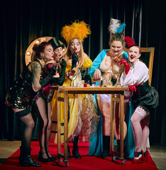 A group of female cabaret performers in diverse vintage costumes gather around a small table laden with makeup and props on a red carpet. Concept of cabaret, show, circus, retro, vintage, performance. © master1305