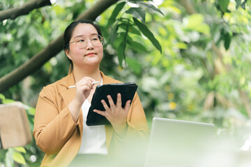 Thoughtful woman in tan blazer uses digital tablet with stylus while sitting outdoors in lush green , surrounded by plants and trees, conveying focus and creativity