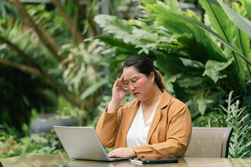 Woman in brown blazer sits at outdoor table surrounded by lush greenery, working on laptop. She...