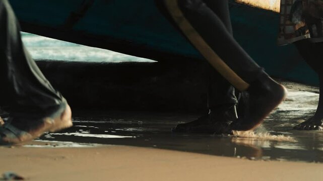 Close-up of local feet pushing colorful Senegalese pirogue onto Cap Skirring beach &mdash; traditional fishing effort, slow shutter motion blur, textured sand, movement, and tropical coastal life scene