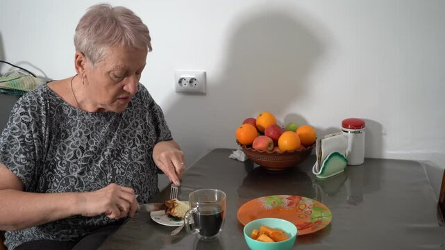 Eat cottage cheese casserole pie. Elderly woman drinking coffee from transparent cup. Grandma having breakfast. Fruit on table in kitchen. Food at home. Fork knife in hands