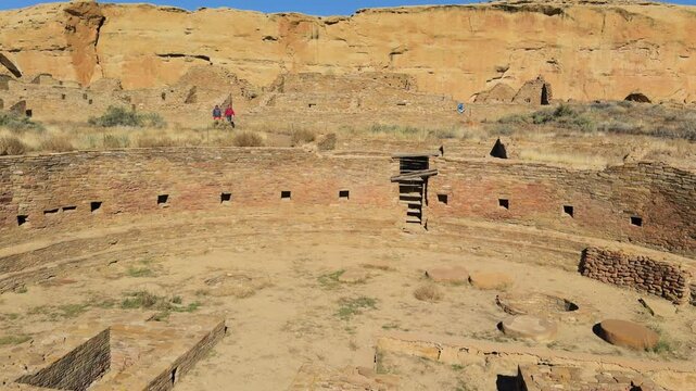Wide view of circular kiva ruins where foreign visitors walking around in desert terrain at Chaco Canyon, New Mexico.