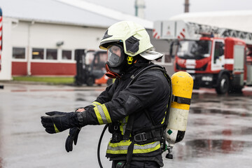 Firefighter in full protective gear adjusts gloves during a rainy day at a fire station. Fire truck...